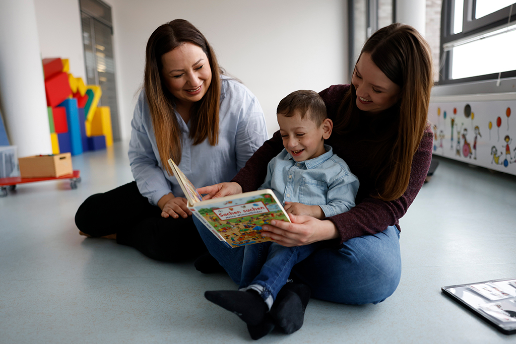 Ein Junge brachtet mit seiner Mutter und einer Mitarbeiterin der Frühförderung ein Bilderbuch.