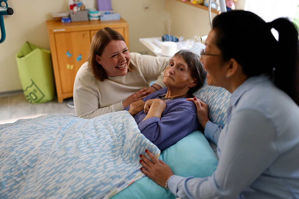 Im Christine-Heuser-Haus: Eine Bewohnerin liegt im Bett - an ihrer Seite sitzen zwei Mitarbeiterinnen.